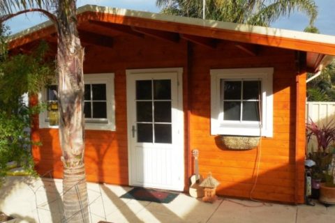 Quaint wooden cabin with white doors and windows, situated in a sunny garden with a palm tree, boasting Shire Council approval. Phoenix Patios, Cottages and Granny Flats in Perth