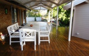 A wooden deck with a white outdoor dining set and a small dog lying on the table under a covered Perth patio. Phoenix Patios, Cottages and Granny Flats in Perth