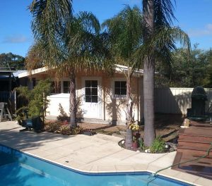 A suburban backyard with a swimming pool, palm trees, and a small single-story house featuring Perth patios in the background on a sunny day. Phoenix Patios, Cottages and Granny Flats in Perth