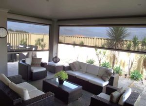 An elegant outdoor living space with modern wicker furniture and outdoor blinds Perth, featuring a landscaped garden visible through large screen windows. Phoenix Patios, Cottages and Granny Flats in Perth