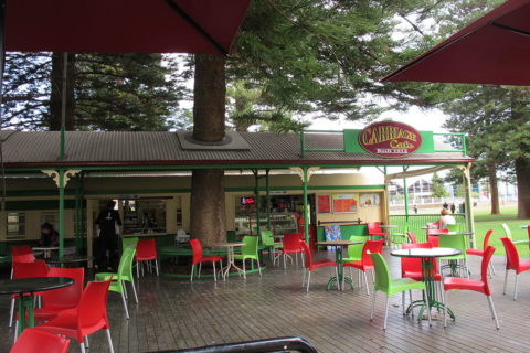 Outdoor cafe with red and green chairs, metal tables, and a canopy. Counters and menu boards are visible in the background, and there is greenery and trees around the seating area.