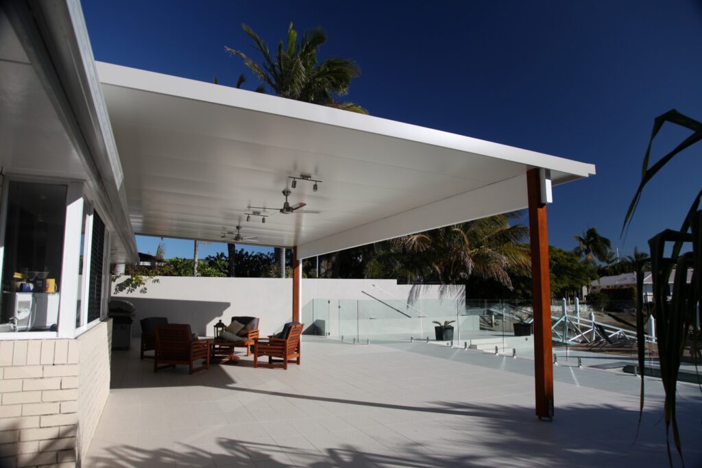 A modern outdoor patio with white flooring, wooden posts, and a roof, featuring a lounge area with chairs and a table. A glass fence encloses the space, with tropical plants in the background.