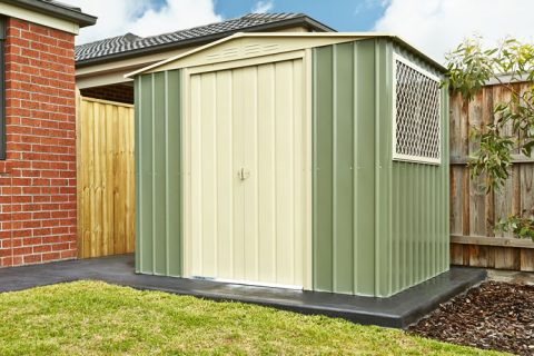 A small, green and beige metal shed with a window on one side is situated next to a brick building and a wooden fence in a backyard.