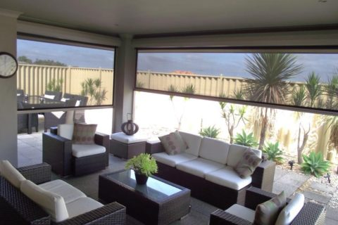 Outdoor patio with wicker furniture and white cushions under a covered area. A glass table sits in the center, and the background features a fence and tall plants.