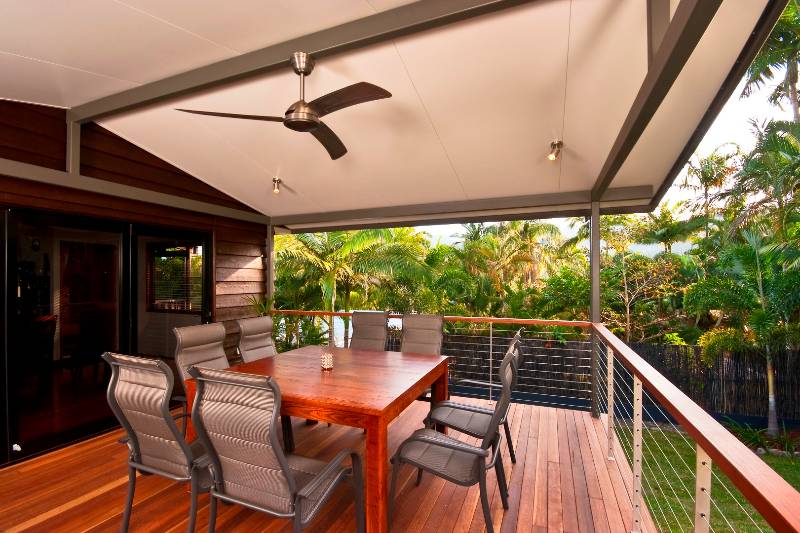 A covered deck with a ceiling fan, wooden dining table, and six chairs. The area is surrounded by a metal railing and a view of lush greenery.